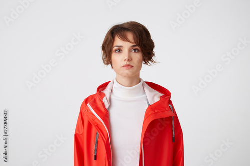 Portrait of young cute short haired girl dressed in white golf and red rain coat, looking at the camera with a calm expression, standing over white background.