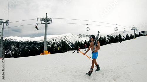 Happy smiling naked man walks on ski slope under lift in slow motion. He wears helmet, goggles and boots only