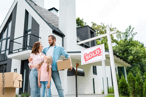 bearded man holding box and standing with wife and daughter near house and board with sold letters