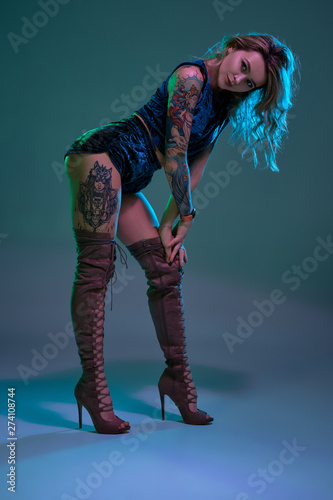 Attractive young blonde girl in blue velour booty shorts and maroon suede high boots posing on camera in studio
