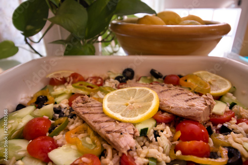 Tasty rice salad with fresh vegetables and grilled mackerel and in the background a bowl with lemons next to green leaves