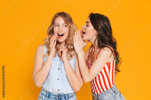 Portrait of two pretty blonde and brunette women 20s in summer wear expressing surprise while whispering gossips