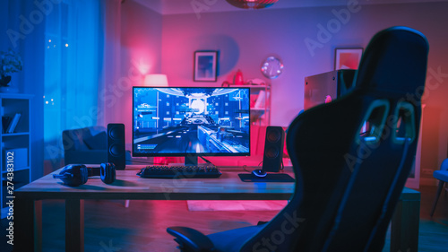 Powerful Personal Computer Gamer Rig with First-Person Shooter Game on Screen. Monitor Stands on the Table at Home. Cozy Room with Modern Design is Lit with Pink Neon Light.