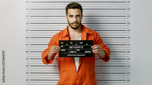 In a Police Station Arrested Man Getting Front-View Mug Shot. He's Wearing Prisoner Orange Jumpsuit and Holds Placard. Height Chart in the Background.