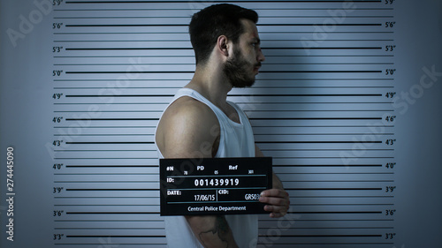 In a Police Station Arrested Beaten Man Poses for Side View Mugshot. He Wears Singlet, is Heavily Bruised and Holds Placard. Height Chart in the Background. Shot with Dark Cold Lights, Vignette Filter