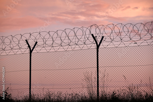 Section of a chainlink fence with razor wire and grass in front of a sunset