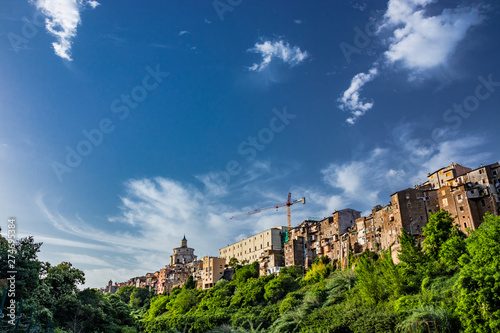 A view of the city of Zagarolo, with the houses built sheer above a tuff hill. Above the roofs, the dome of the church of San Pietro and a crane emerge. The valley full of trees. Rome, Lazio