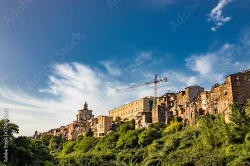 A view of the city of Zagarolo, with the houses built sheer above a tuff hill. Above the roofs, the dome of the church of San Pietro and a crane emerge. The valley full of trees. Rome, Lazio