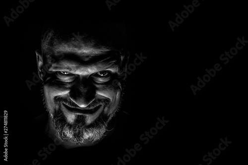  Face with a bearded man grimace against a dark background with sharp shadows. Comedic, fabulous villain or negative character conception with copy space.