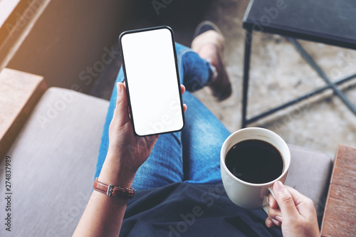 Top view mockup image of woman holding black mobile phone with blank screen while sitting and drinking coffee in cafe