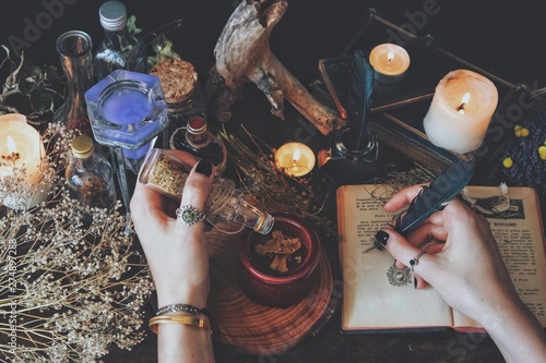 A witch casting a spell and writing it down in her Book of Shadows with a green black feather and ink. Wiccan witch altar filled with nature objects branches dried flowers herbs burning candles potion