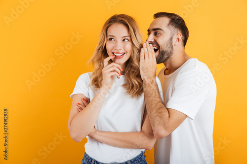 Image of happy couple man and woman in basic t-shirts whispering secrets or gossips to each other