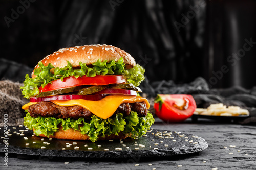 Beef burger with cheese, tomatoes, red onions, cucumber and lettuce on black slate over dark background. Unhealthy food
