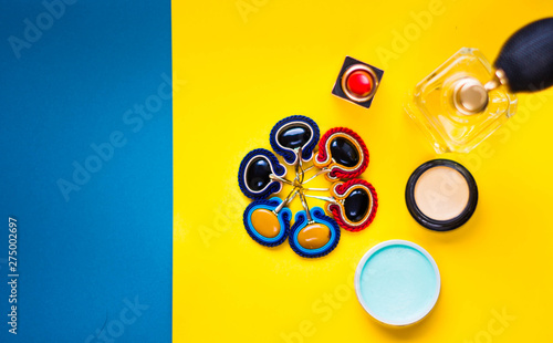 Modny kolaż zawierający ręcznie szyte kolczyki sutasz, szminkę, perfumy oraz kolorowe kosmetyki w ujęciu top view na żółto niebieskim tle