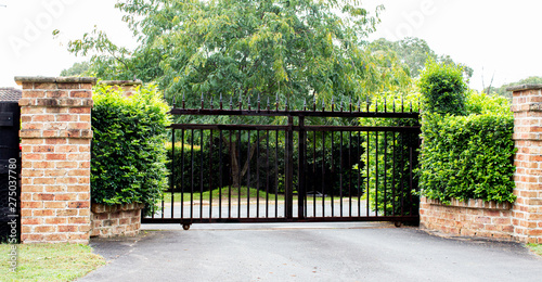Black metal driveway property entrance gates set in brick fence with garden shrubs and trees in background