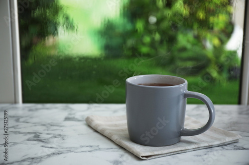 Cup of hot drink and napkin on marble windowsill against glass with rain drops, space for text