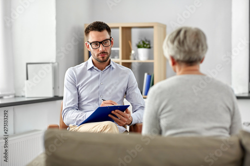 geriatric psychology, mental therapy and old age concept - psychologist with clipboard taking notes and listening to senior woman patient at psychotherapy session