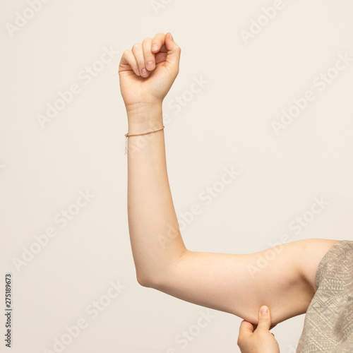A closeup view of a young Caucasian lady squeezing her upper arm, showing the sagging skin and fat beneath the triceps, commonly referred to as bingo wings. Isolated against a white background.