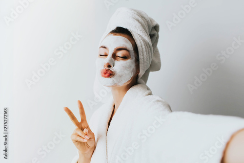 Self portrait of charming, stylish, pretty, model after bath wrapped in towel shooting selfie on front camera applying using face mask for her dry, oiled, sensual face skin, enjoying procedure