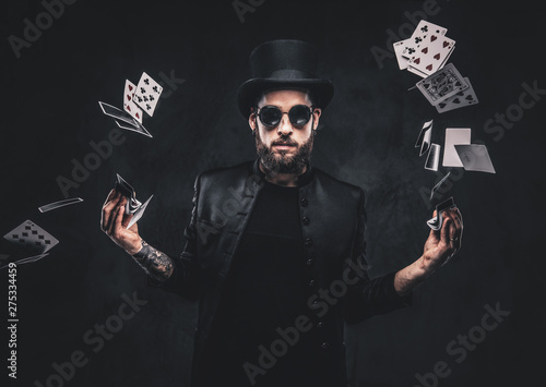 Magician in a black suit, sunglasses and top hat, showing trick with playing cards on a dark background.