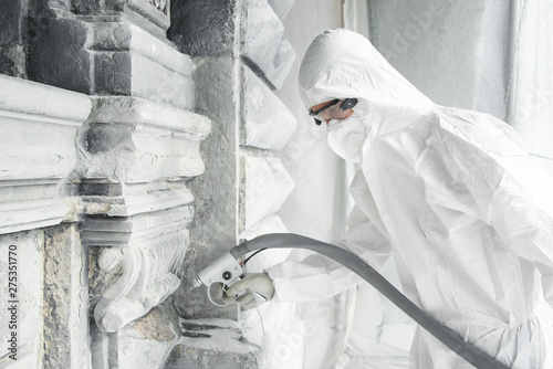 Man in a white protective uniform cleans stone carved sculpture from the dirt and concrete with a sandblasting machine. Restoration of stone sculpture. A jet of sand under high pressure.