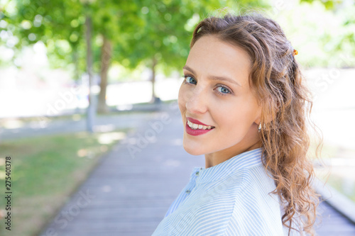Relaxed happy girl enjoying nature in park. Headshot of young woman with wavy hair looking at camera and smiling. Pretty woman outdoors concept