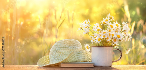 beautiful composition with chamomile flowers in Cup, old book, braided hat in summer garden. Rural landscape natural background with Chamomile in sunlight. Summertime season. copy space