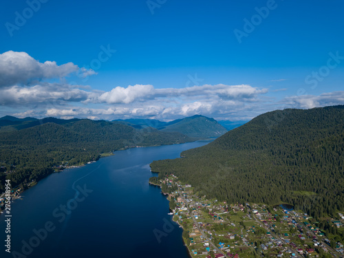 Aerial view of mouth at wide Teletskoye lake in the Altai Mountains by the blue water, sky with white clouds, green trees on the slopes of the rocks and village on shore. Rest and travel in nature.