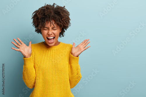 Dissatisfied angry curly woman expresses hate and rage, screams loudly, raises palms, keeps mouth widely opened, dressed in yellow knitted sweater, disagrees with somebodys opinion, being irritated