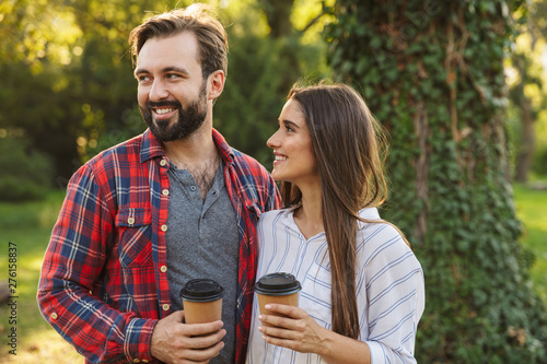 Cheerful young loving couple walking outdoors in a green nature park forest drinking coffee.