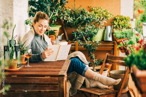 Young beautiful woman relaxing on cozy balcony, reading a book, wearing warm knitted pullover, glass of wine on wooden table
