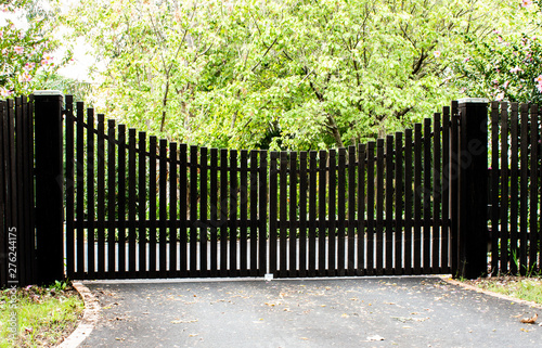 Dark wooden driveway property entrance gates set in timber picket fence with garden shrubs and trees in background