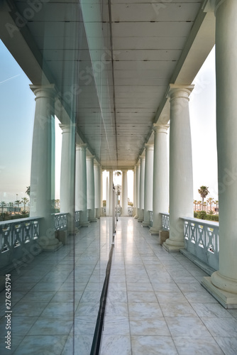 Exterior corridor overlooking the sea of a building with classic white columns reflected in a glass.