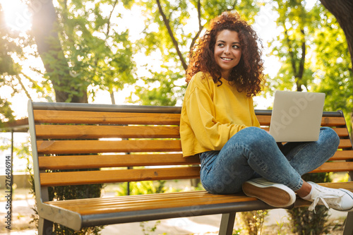 Happy young beautiful curly student girl sitting outdoors in nature park using laptop computer.