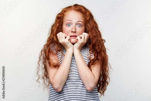 Portrait of afraid little girl with ginger hair and freckles, scared and anxious biting her finger nails, looking at camera with wide opened eyes and looks away, isolated over white wall.