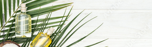 top view of coconut oil in bottles on green palm leaf on white wooden surface, panoramic shot