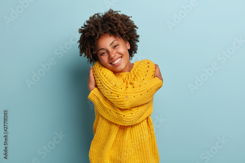 Lovely happy African American woman daydreams, embraces herself, recalls romantic date, feels comfort, keeps hands across body sensually, has broad smile, wears yellow sweater, poses indoor.