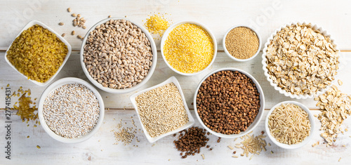 Selection of whole grains in white bowls - rice, oats, buckwheat, bulgur, porridge, barley, quinoa, amaranth, on white wood background