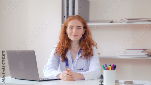 Portrait redhead doctor sitting at the desk in office. Candid professional woman therapist posing looking at the camera and friendly smiling.