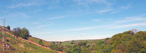 a long panoramic view of west yorkshire countryside with fields and moors above the woodland in hardcastle crags with a blue sunlit cloud sky