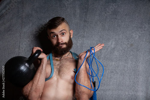 The guy chooses between a jump rope and free weights. Concept cardio or strength. Portrait of a sporty, bearded guy with sport shells, in the studio on a gray background.