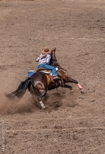 Barrel Racing at a Rodeo, cowgirl riding a dark colored horse around a barrel. She has a pony tail and is wearing a large tan cowboy hat. The dirt is flying as the horse digs in.