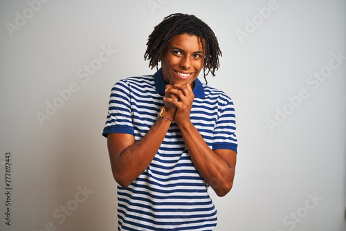 Afro man with dreadlocks wearing striped blue polo standing over isolated white background laughing nervous and excited with hands on chin looking to the side