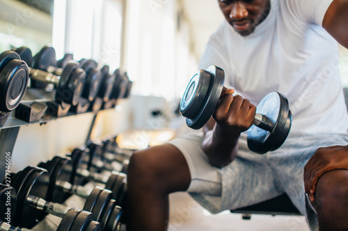 Young African American man sitting and lifting a dumbbell close to the rack at gym. Male weight training person doing a biceps curl in fitness center