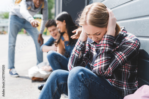 selective focus of smiling friends bulling and pointing with fingers at sad blonde teen