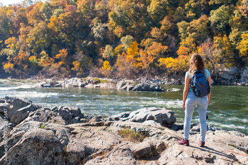 Young woman photographer with backpack and camera looking at view of Potomac river in Great Falls with autumn colorful foliage in Maryland