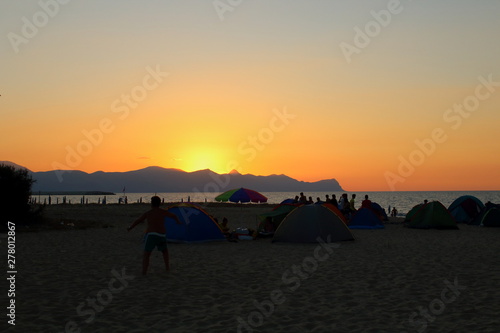evocative image of sunset over the sea promontory in the background and silhouette of tents and people camping on the beach 