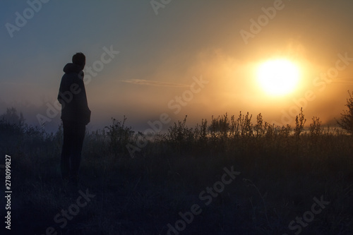 Silhouette of the traveler against fog landscape over a flower meadow, the first rays of dawn and dark silhouettes of trees against a sunrise, selective focus