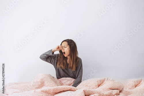 The girl is in bed and yawns. She covers her mouth with her hand. Early morning and she is sleepy.