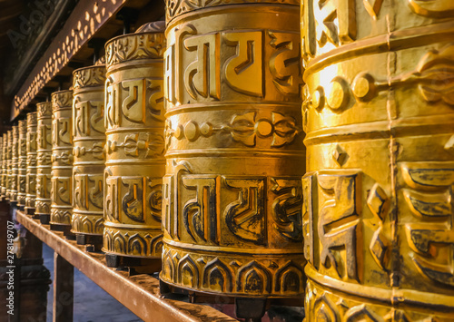 A prayer wheels on a spindle made from metal and wood. Mantra Om Mani Padme Hum is written in Newari language of Nepal on the outside of the wheels. Golden Temple (Kwa Bahal) buddhist temple in Patan